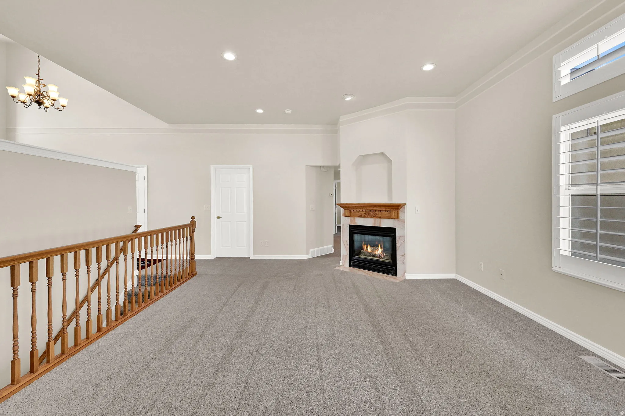 Spacious room featuring gray carpet, a corner fireplace with a wooden mantel and stone surround, and windows with white plantation shutters