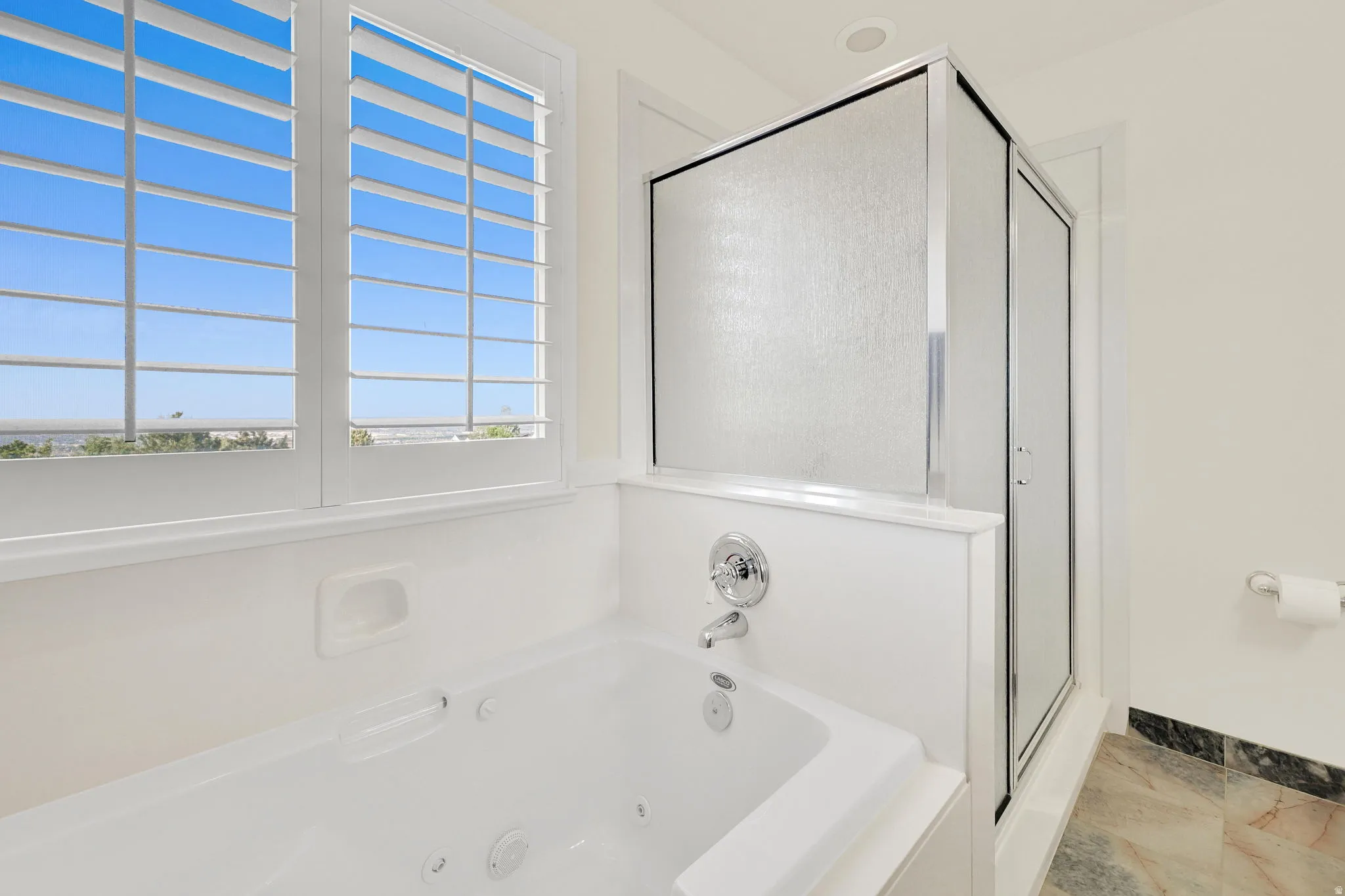 White jetted soaking tub with chrome fixtures, plantation shutters, and an enclosed standing shower with frosted glass panels