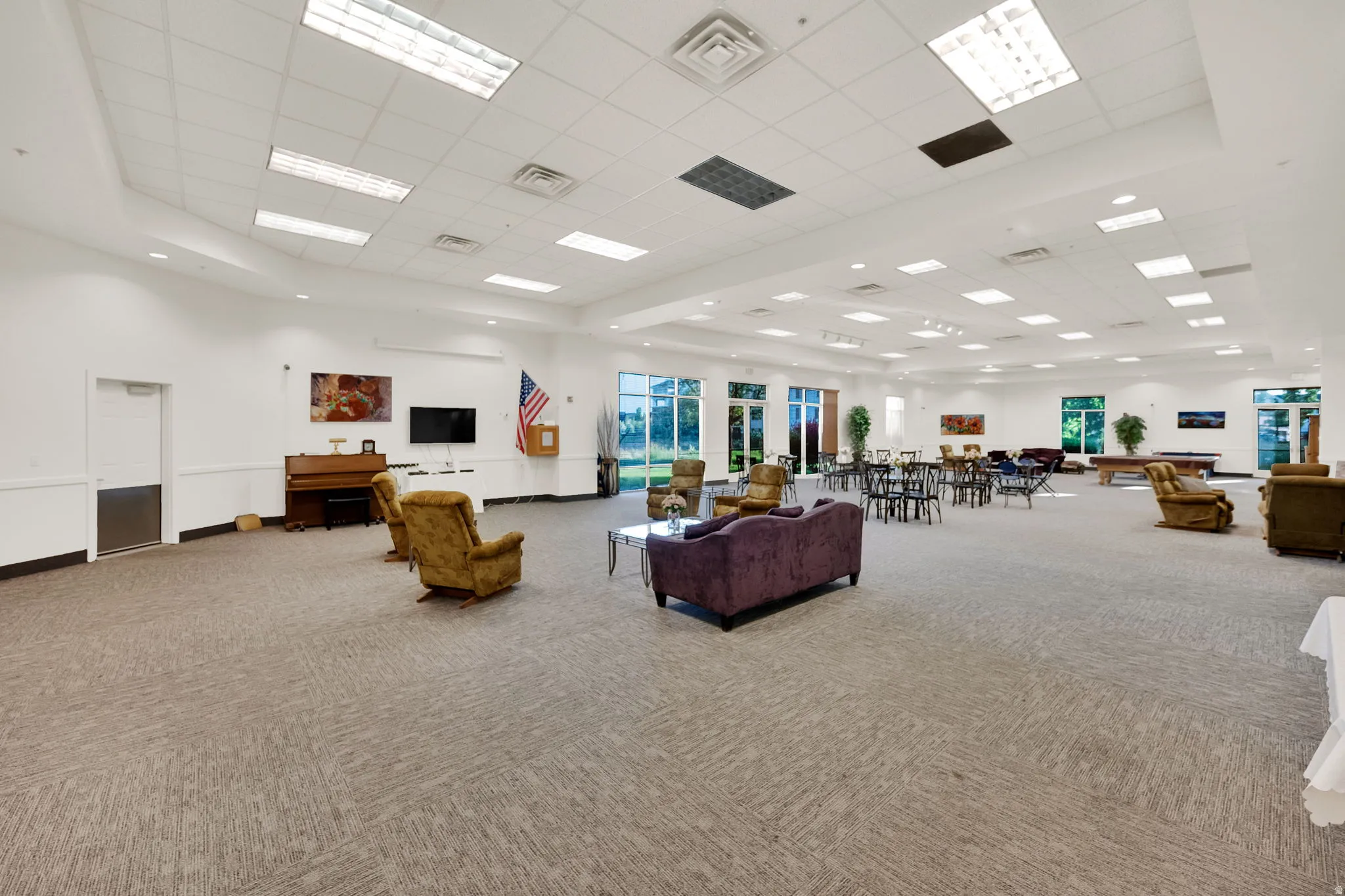 Spacious common room featuring recessed lighting, large windows, neutral patterned carpeting, and white walls with wainscoting
