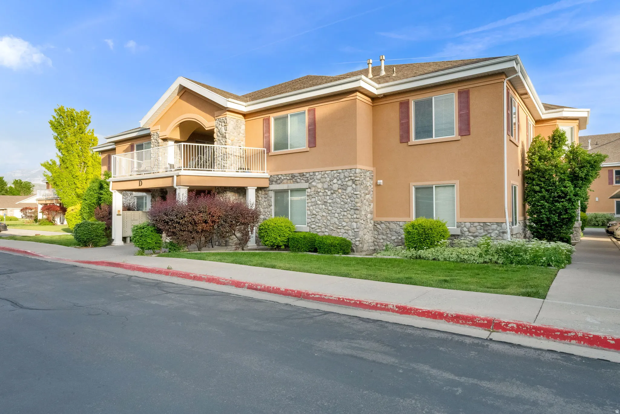 Exterior facade featuring stone accents, stucco finish, a second-story balcony with white railing, red decorative shutters, and mature landscaping