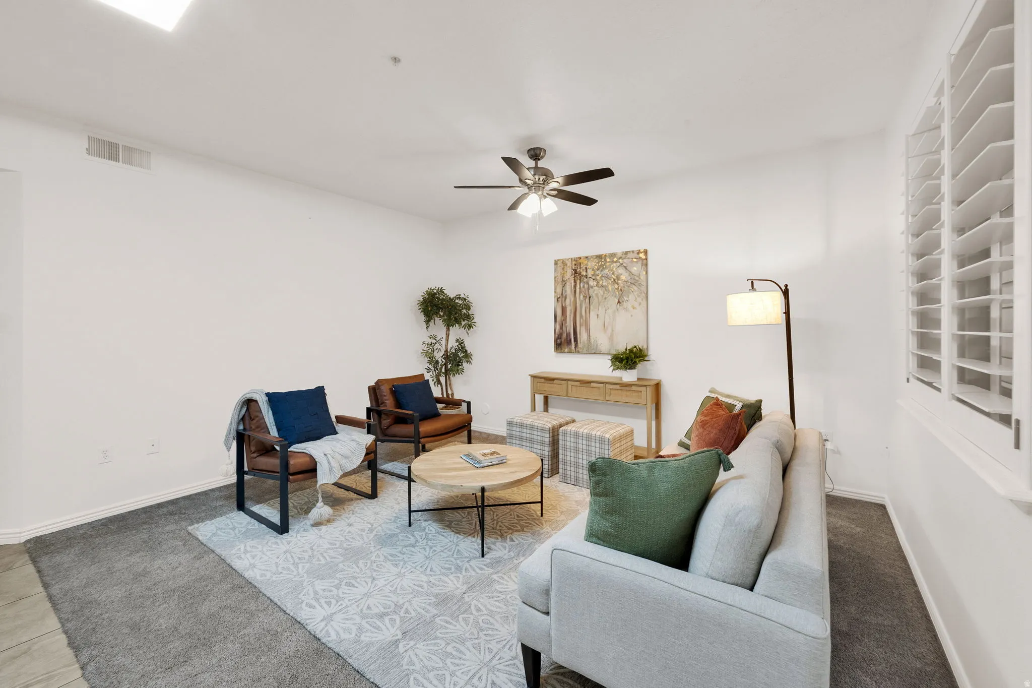 Spacious living area featuring carpet flooring, crisp white walls, ceiling fan with integrated lighting, and plantation-style window shutters