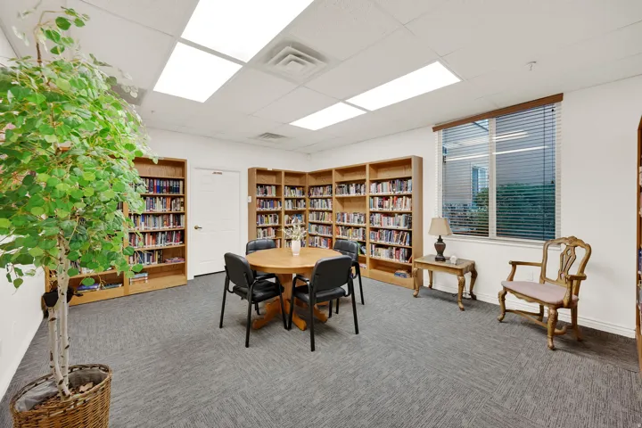 Space featuring extensive built-in wood shelving, grey patterned carpeting, large window with blinds, and white walls