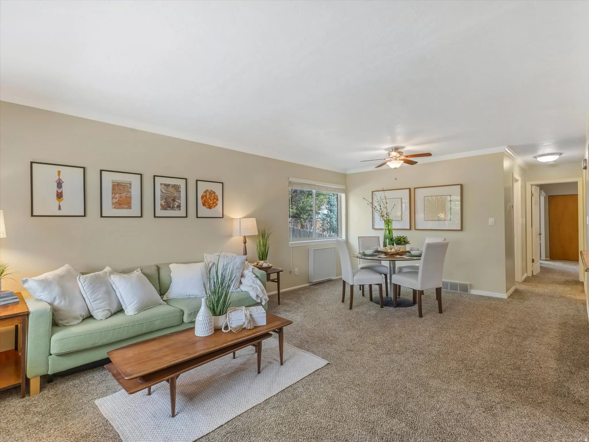 Open-concept living and dining area featuring neutral carpeting, crown molding, and a ceiling fan