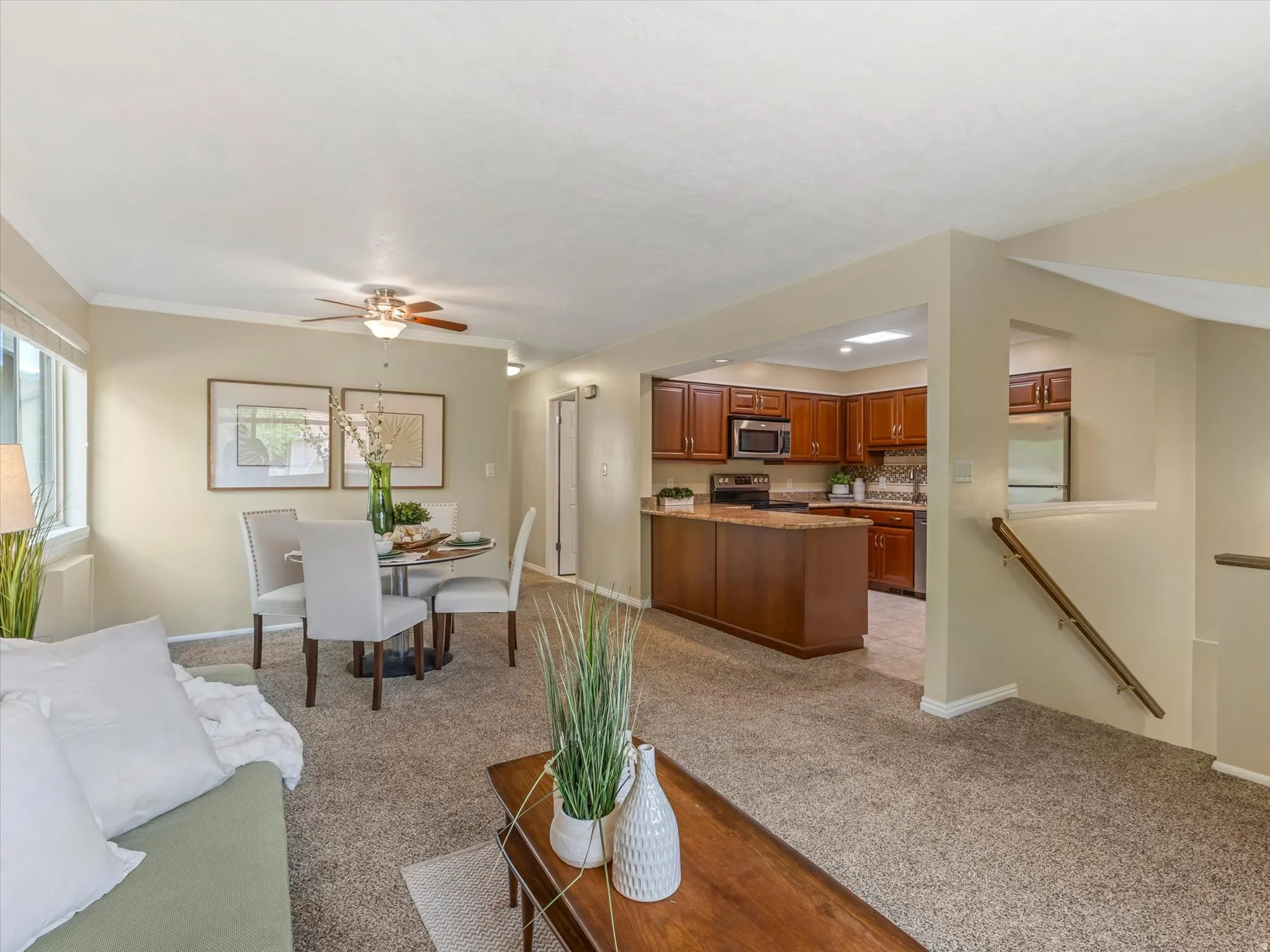 Open-concept living area featuring soft carpet flooring, a dining space with a ceiling fan, and a kitchen with wood cabinetry, granite-finish countertops, and stainless steel appliances