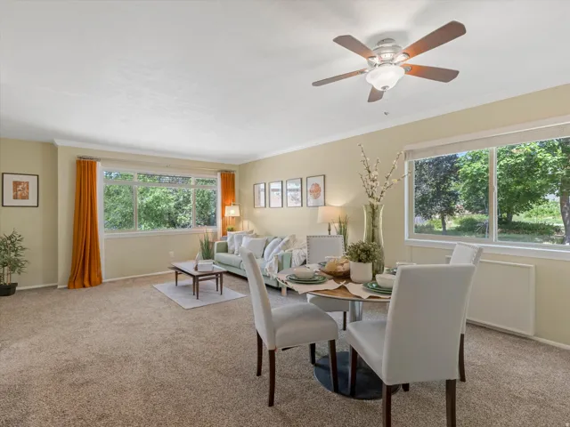 Carpeted living and dining area featuring multiple large windows, crown molding, and a ceiling fan with integrated lighting