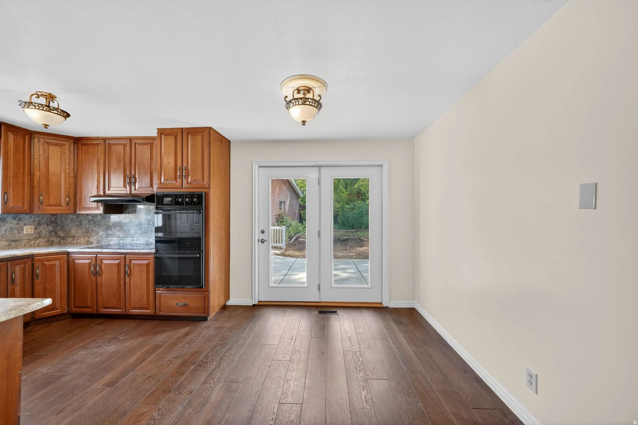 Kitchen area featuring wood-finish flooring, corner cabinetry, integrated double wall ovens, a coordinating range hood, and a light-colored backsplash