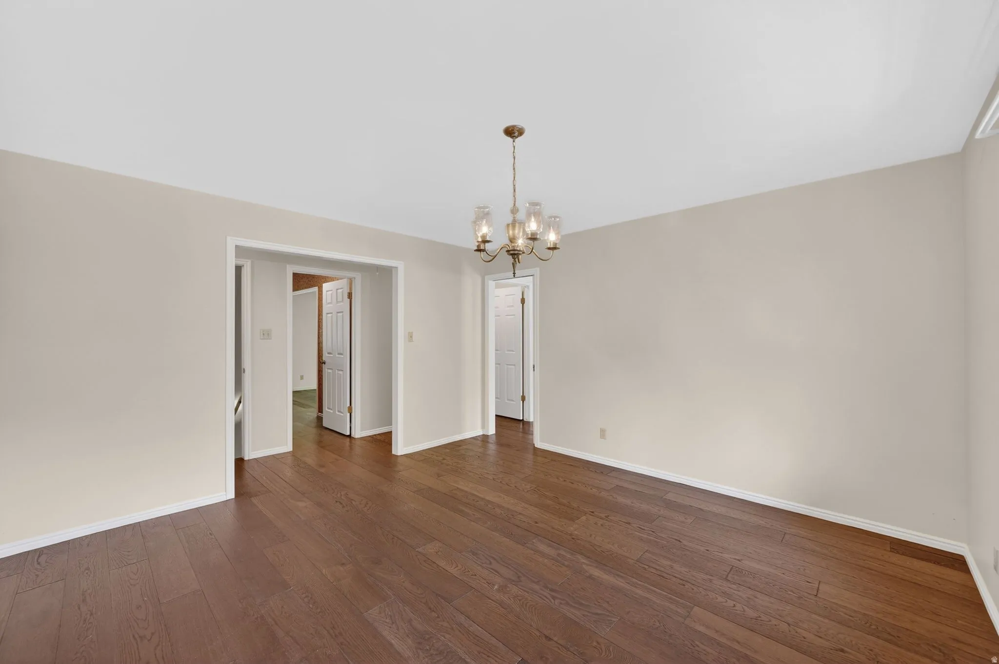 Spacious room featuring wood-finish flooring, a brass-toned chandelier with glass shades, neutral wall paint, and white trim throughout