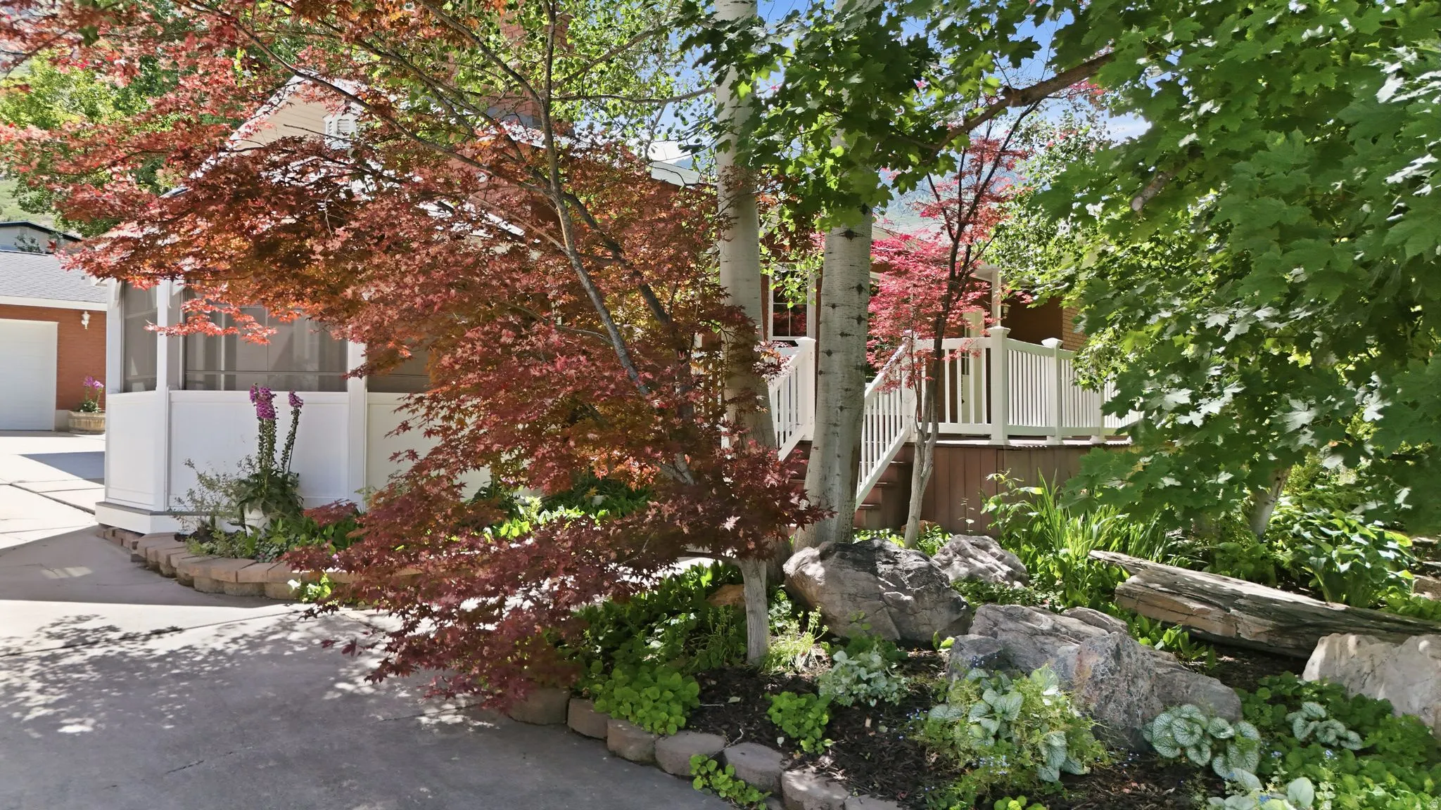 Landscaped front exterior featuring a screened porch, a red Japanese maple, mature deciduous trees, a rock garden, and a paved driveway leading to a garage with a white door