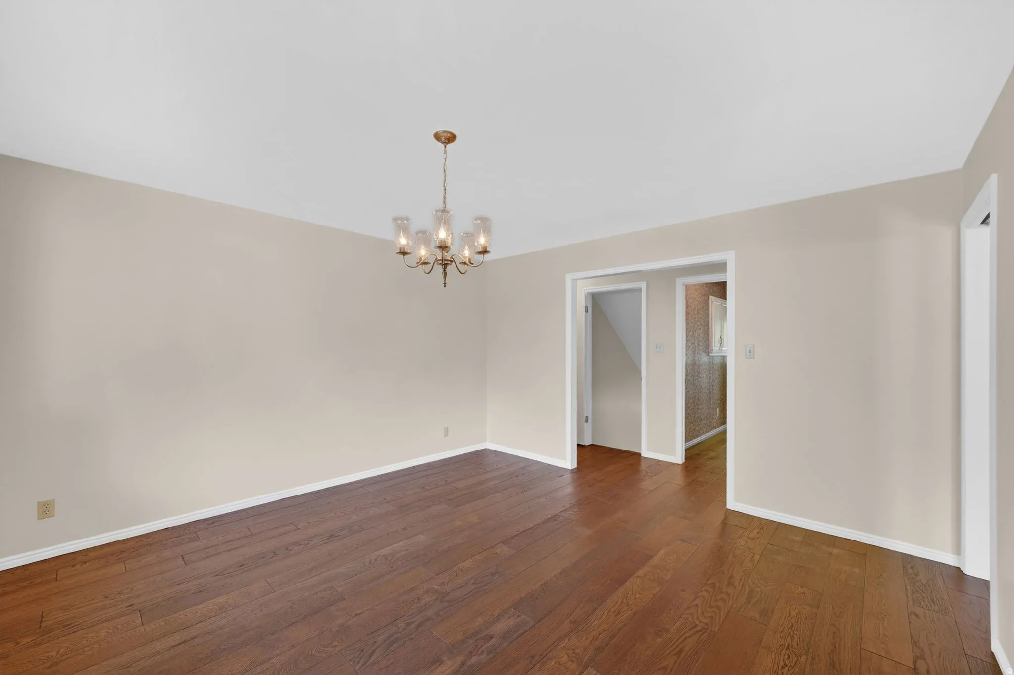 Spacious room featuring wood-finish flooring, a brass chandelier with glass shades, light-toned walls, and white baseboards