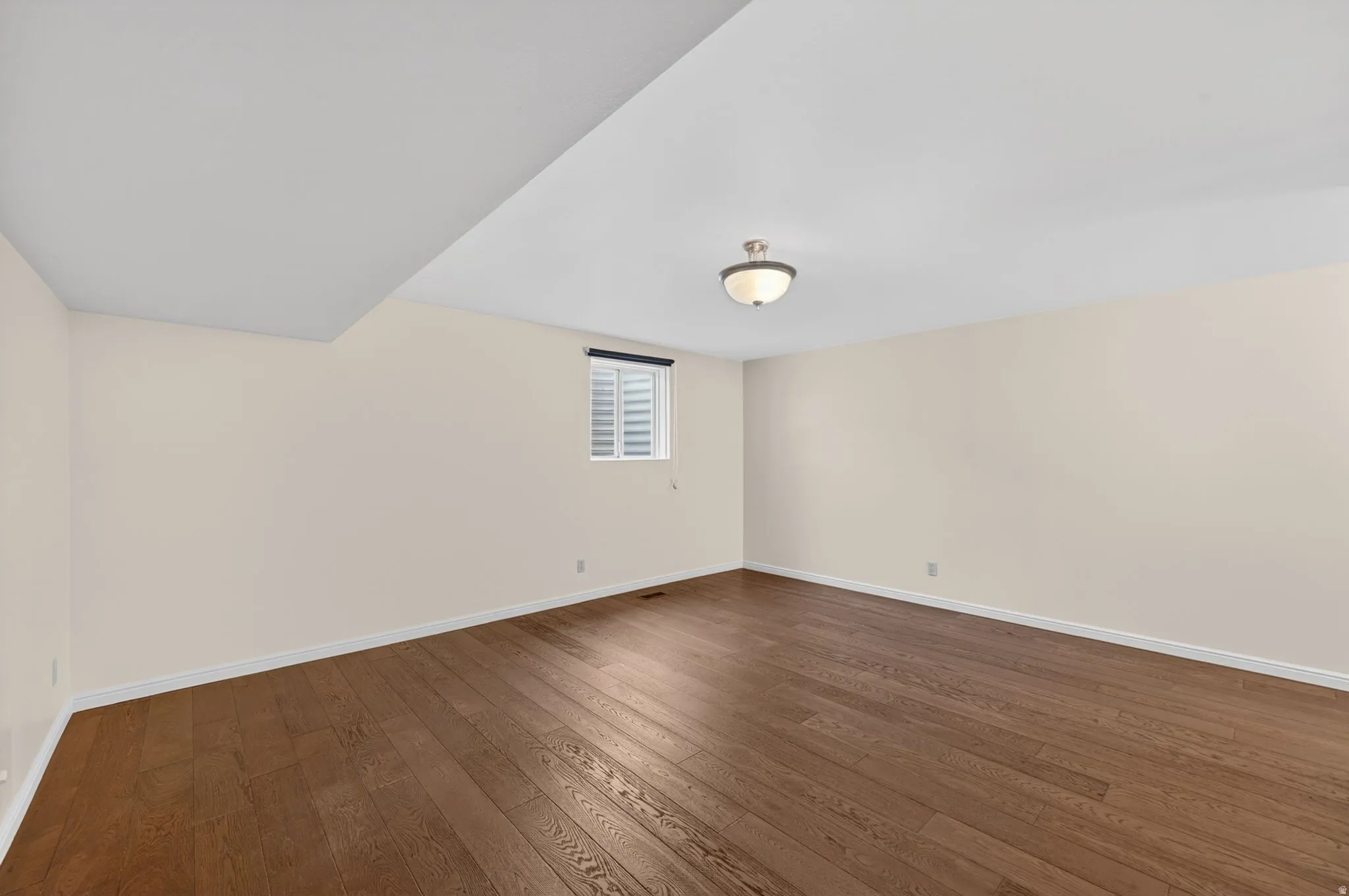 Spacious interior room featuring wood-finish flooring, neutral wall tones, a ceiling-mounted light fixture, and a window with louvered blinds