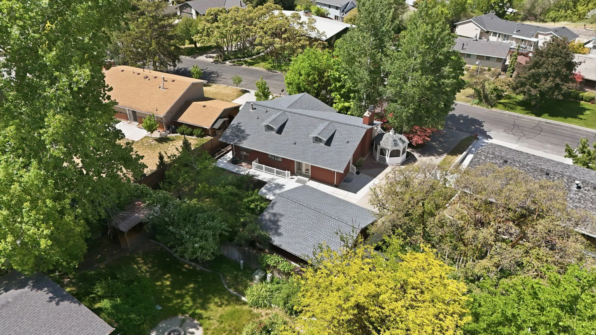 Red brick exterior residence featuring a dark shingle roof with multiple dormer windows, an attached white gazebo, paved patio areas, and mature trees surrounding the property