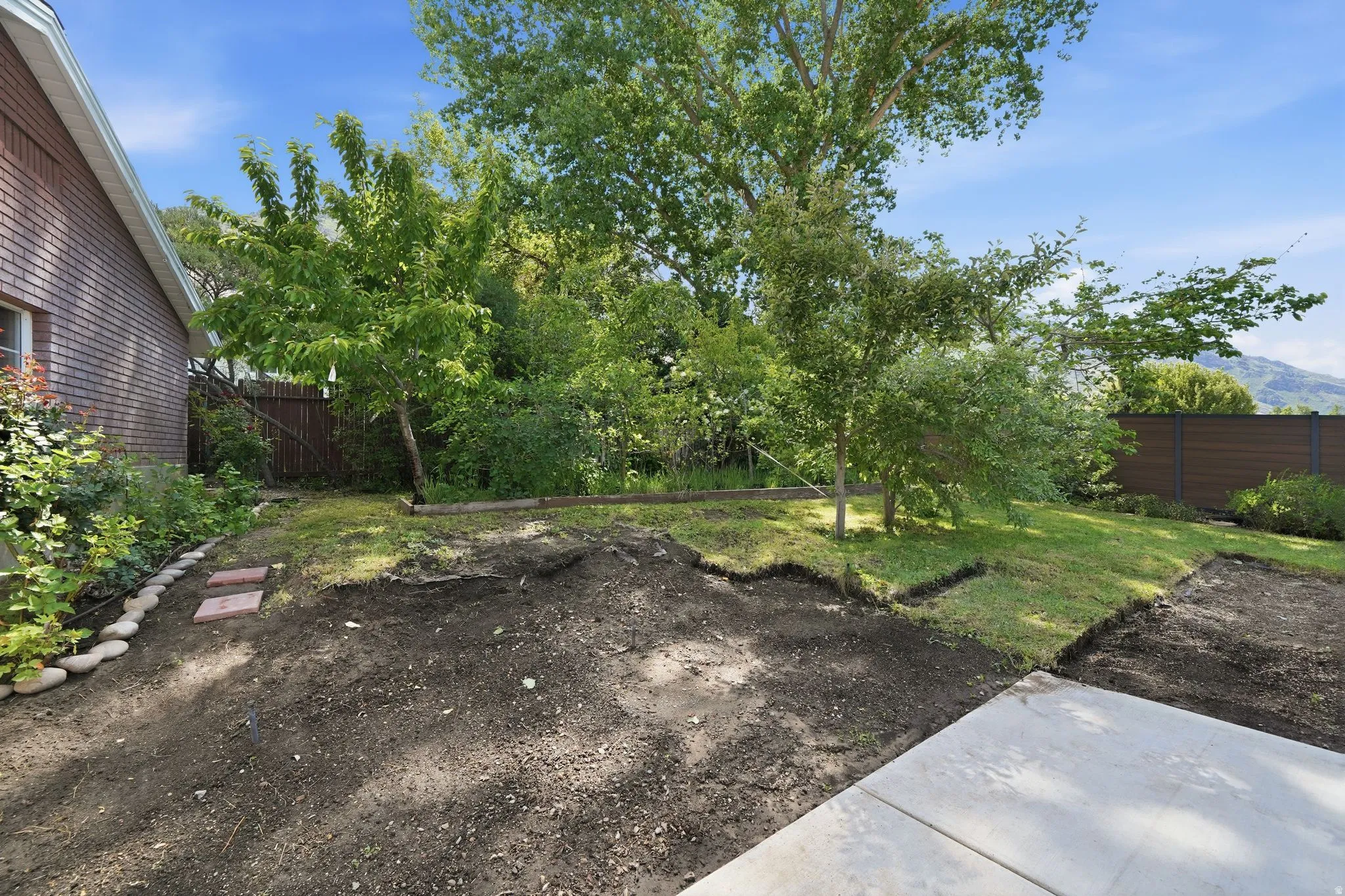 Expansive backyard featuring a partial concrete patio, areas of established lawn, mature trees, a low privacy fence, and brick exterior elements