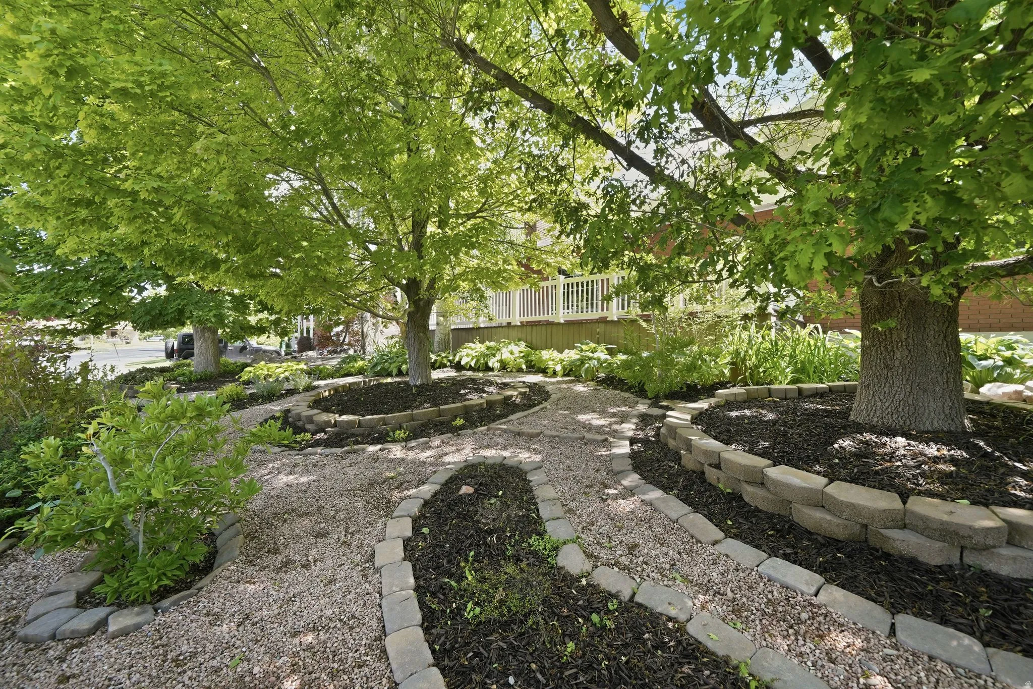 Landscaped exterior featuring a gravel pathway with paver edging, mature trees, and a tiered garden bed with dark mulch