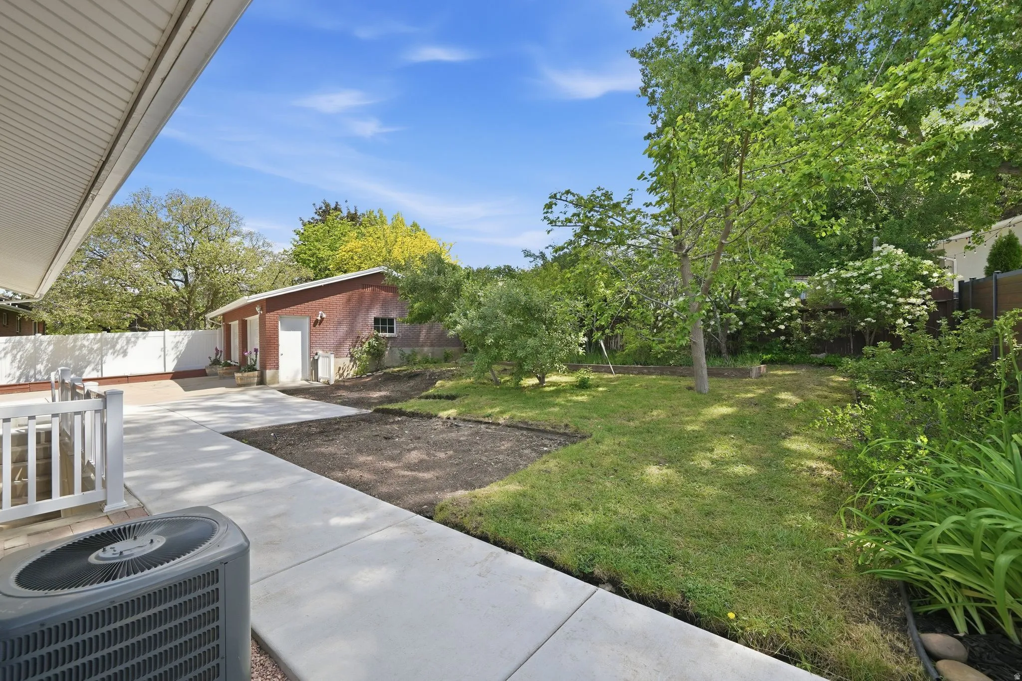Spacious concrete patio and walkways extending to a detached brick outbuilding with multiple doors, surrounded by established trees and green landscaping