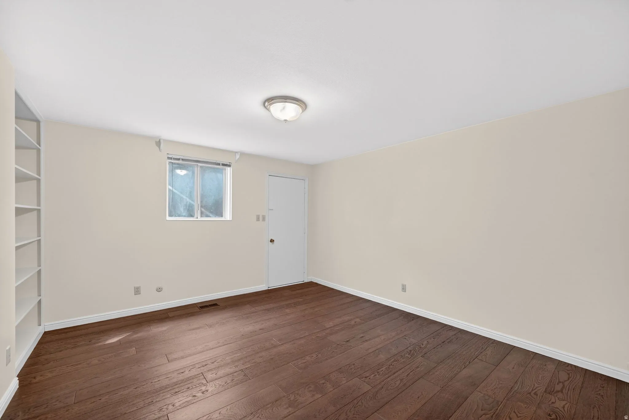 Spacious room featuring rich wood-finish flooring, light neutral walls, and a built-in shelving unit
