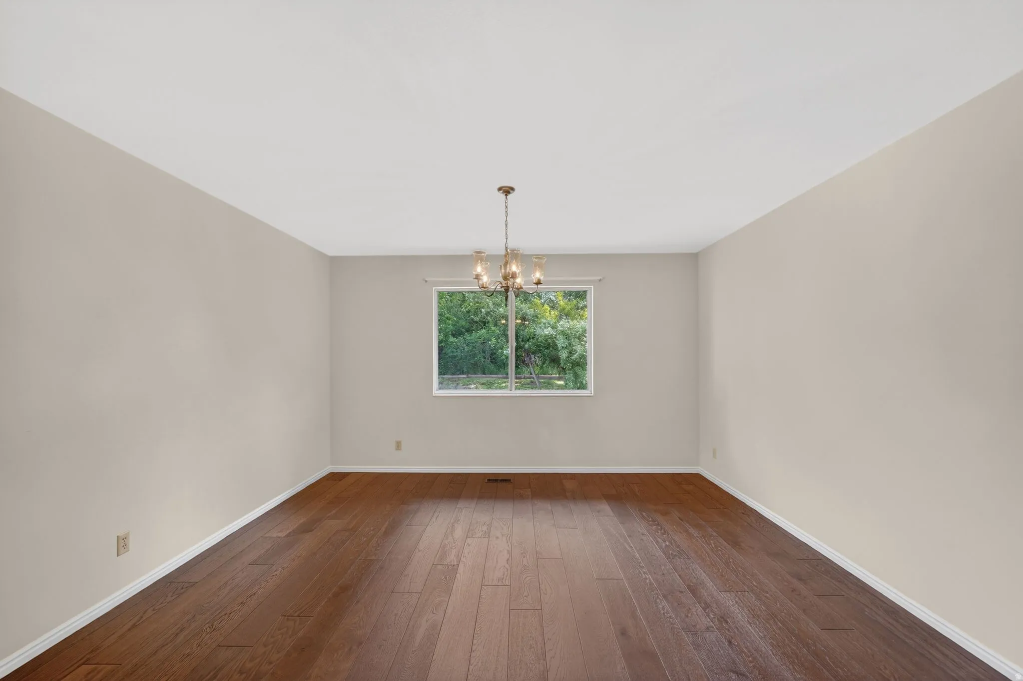 Spacious room featuring rich wood-finish flooring, a contemporary window, white baseboards, light neutral wall tones, and a bronze-finish chandelier