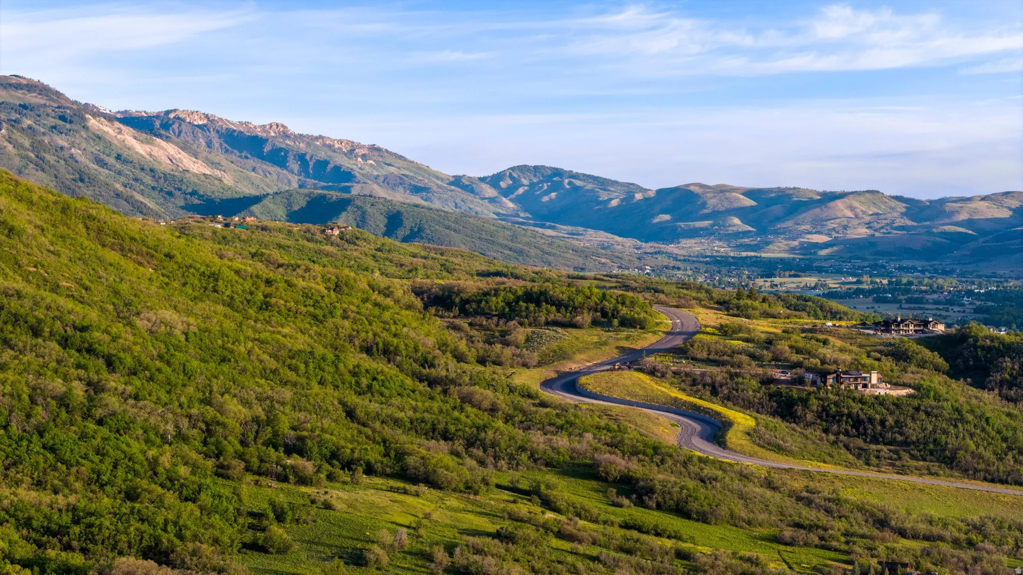 Winding asphalt road through verdant hillsides, featuring distant mountain ranges and scattered residential structures
