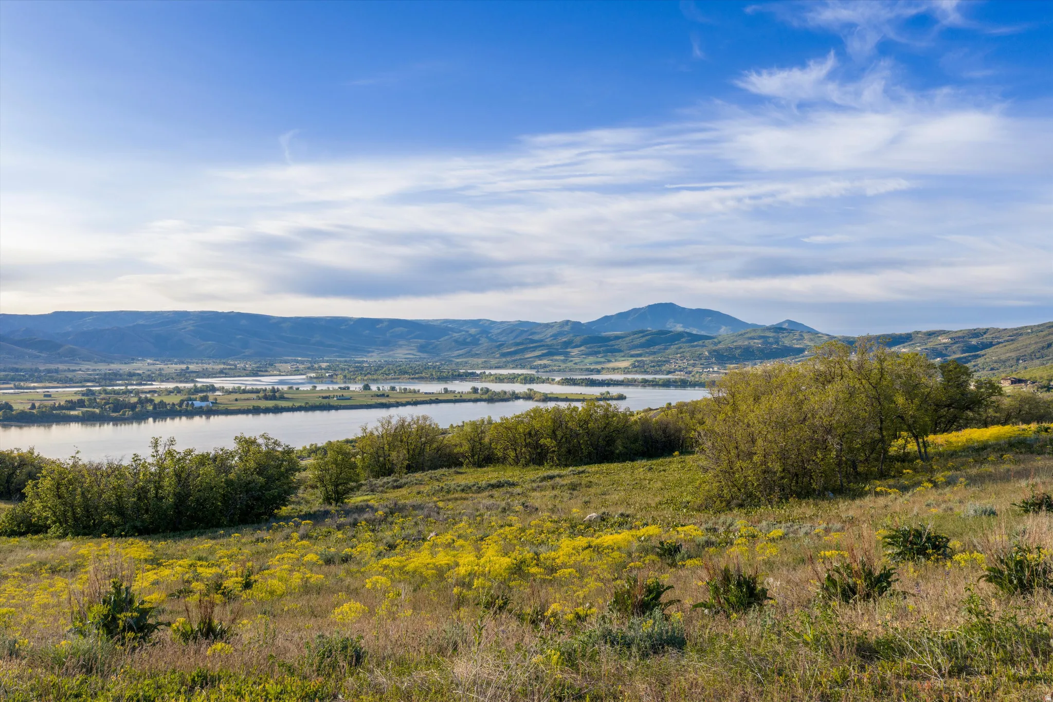 Expansive natural landscape featuring rolling hills with green and yellow foliage, a prominent body of water, distant mountains, and a clear blue sky