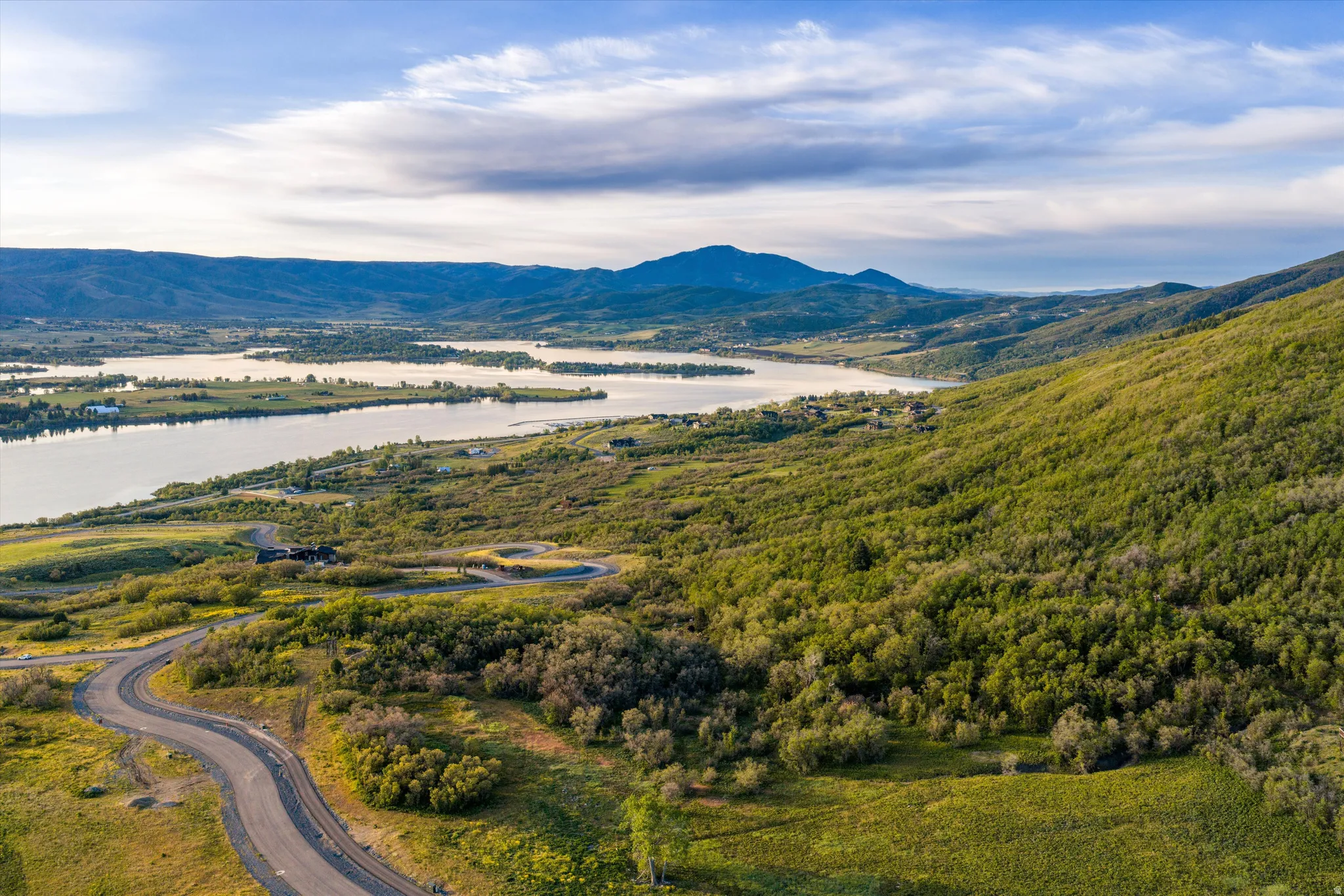 Expansive natural landscape featuring a winding paved road, lush green hillsides, a large body of water, and distant mountains