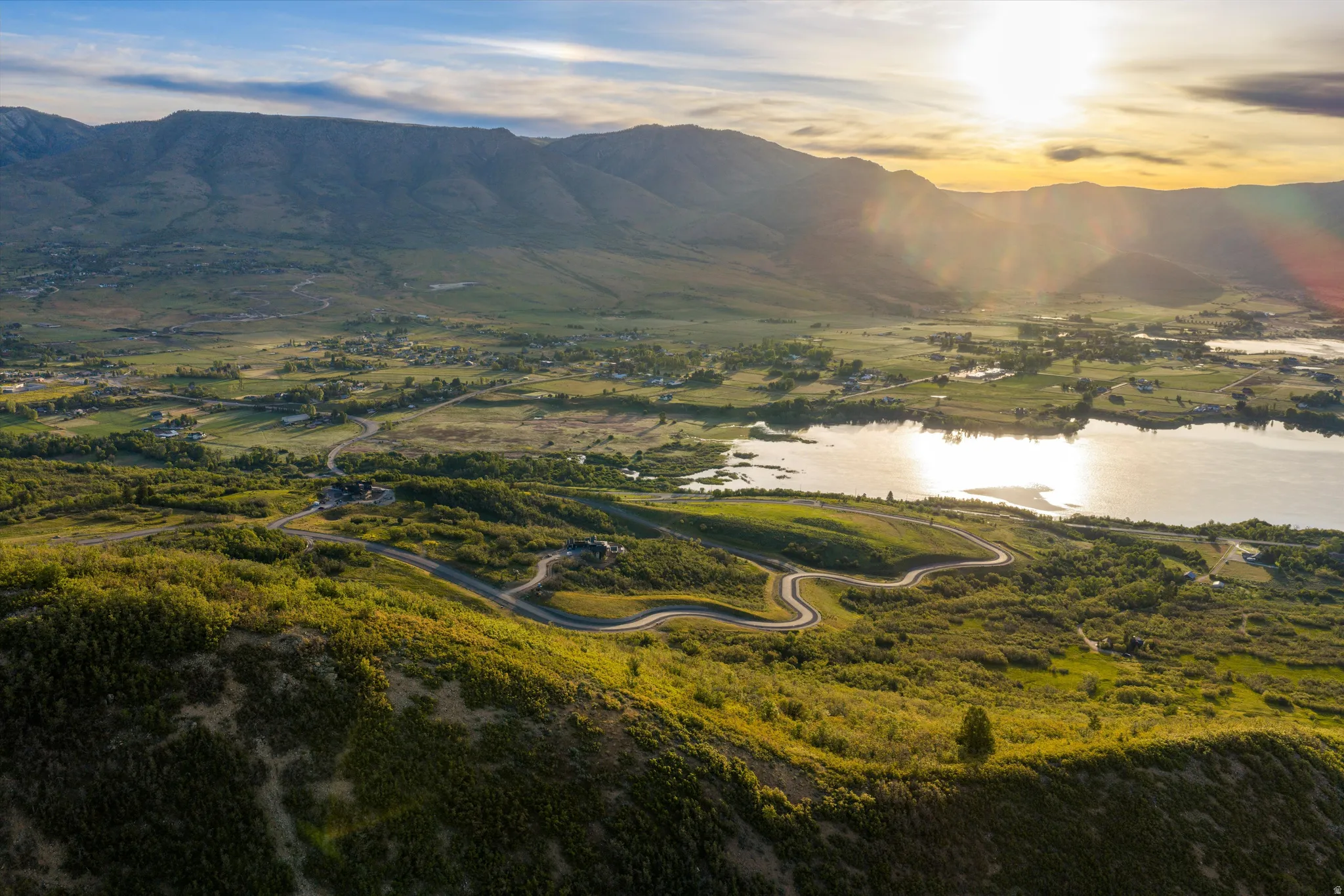 Expansive mountain setting featuring a winding paved road, lush green hillsides, a sparkling lake, and distant valley development