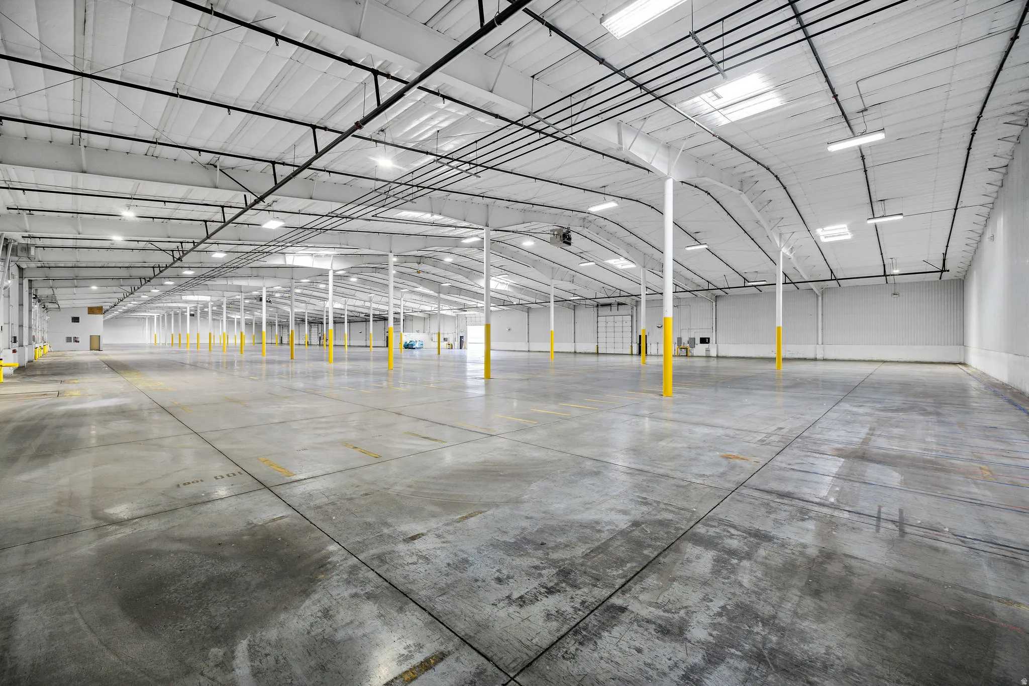 Expansive commercial interior featuring concrete flooring, numerous support columns, exposed ceiling trusses, abundant overhead lighting, and multiple skylights