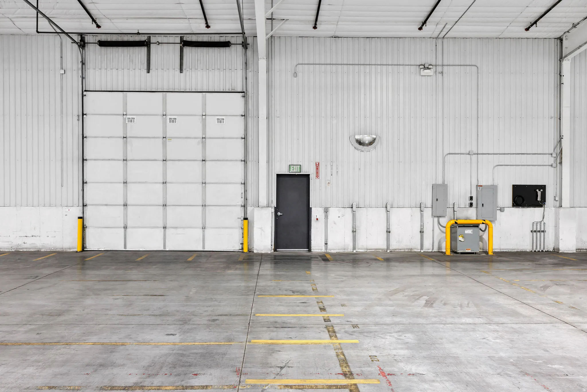 Interior industrial space featuring a large roll-up door with yellow protective bollards, a separate exit door, exposed conduit, and concrete flooring with painted markings
