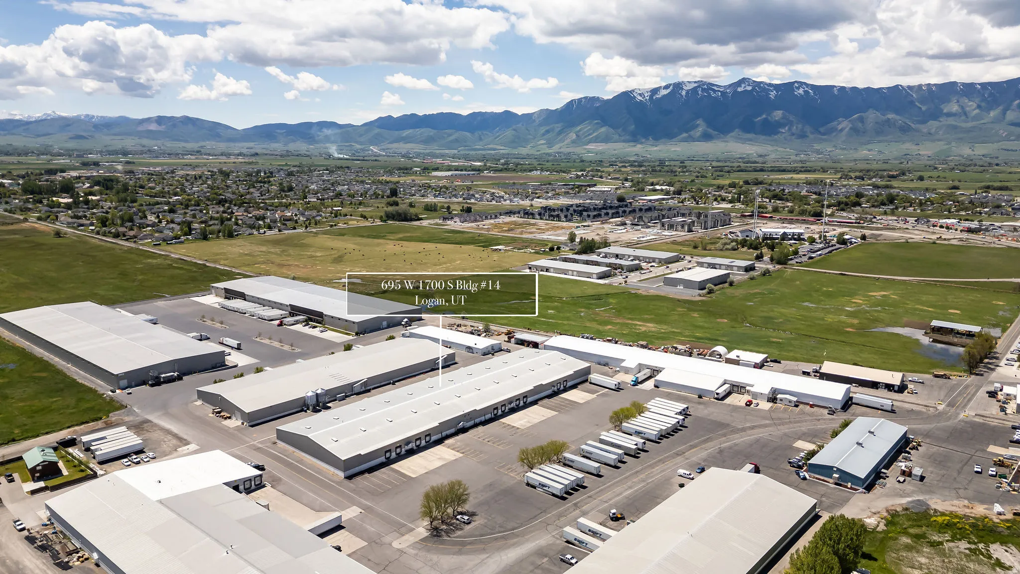 Expansive commercial property featuring multiple large industrial buildings with metal roofs and extensive paved parking areas, situated in a developed commercial zone with visible mountain ranges in the distance