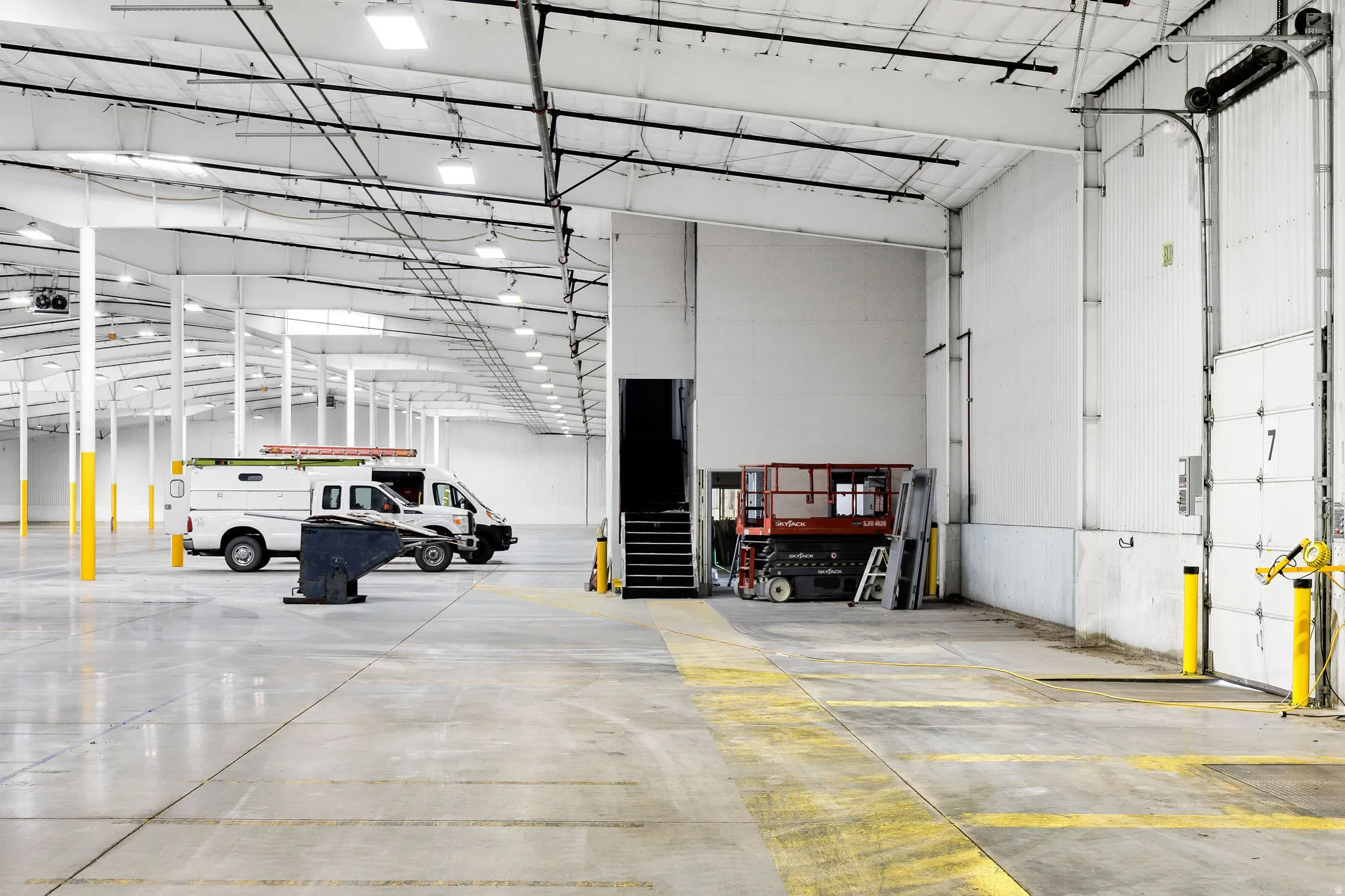 Expansive industrial interior featuring a polished concrete floor, high ceilings with exposed structural beams, numerous overhead light fixtures, multiple support columns, and a large roll-up door with loading dock access