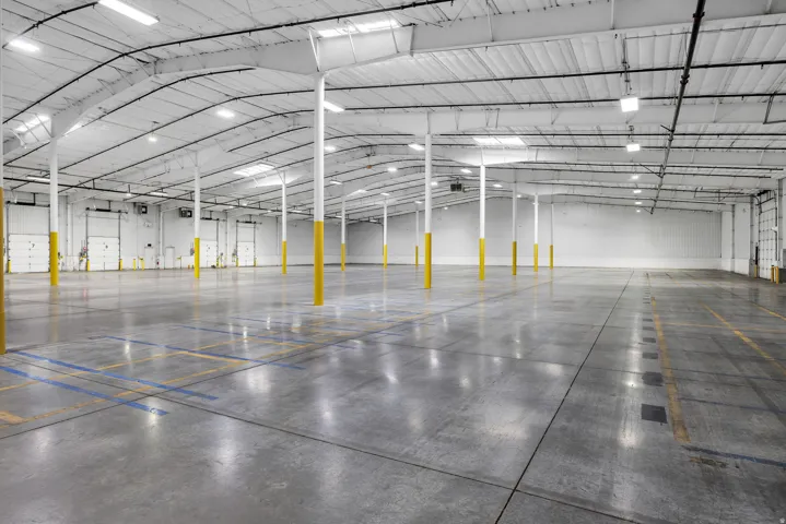 Spacious warehouse interior featuring a high arched ceiling with exposed metal trusses, multiple skylights, and fluorescent lighting