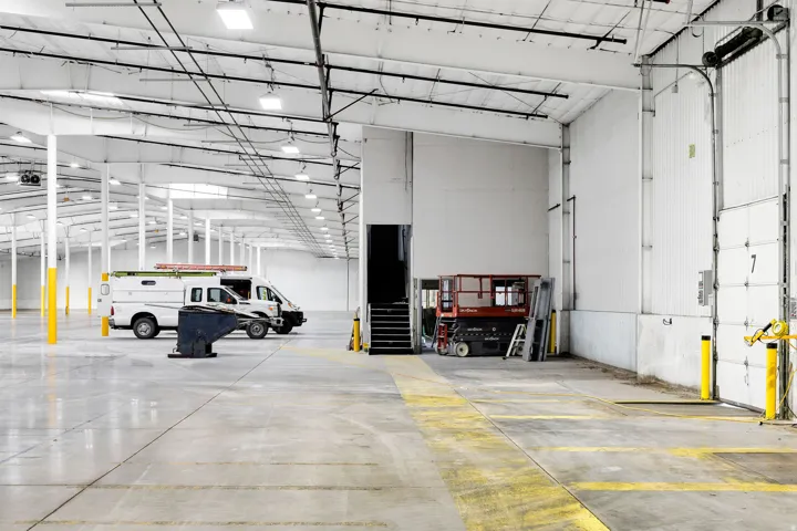 Expansive industrial interior featuring a polished concrete floor, high ceilings with exposed structural beams, numerous overhead light fixtures, multiple support columns, and a large roll-up door with loading dock access