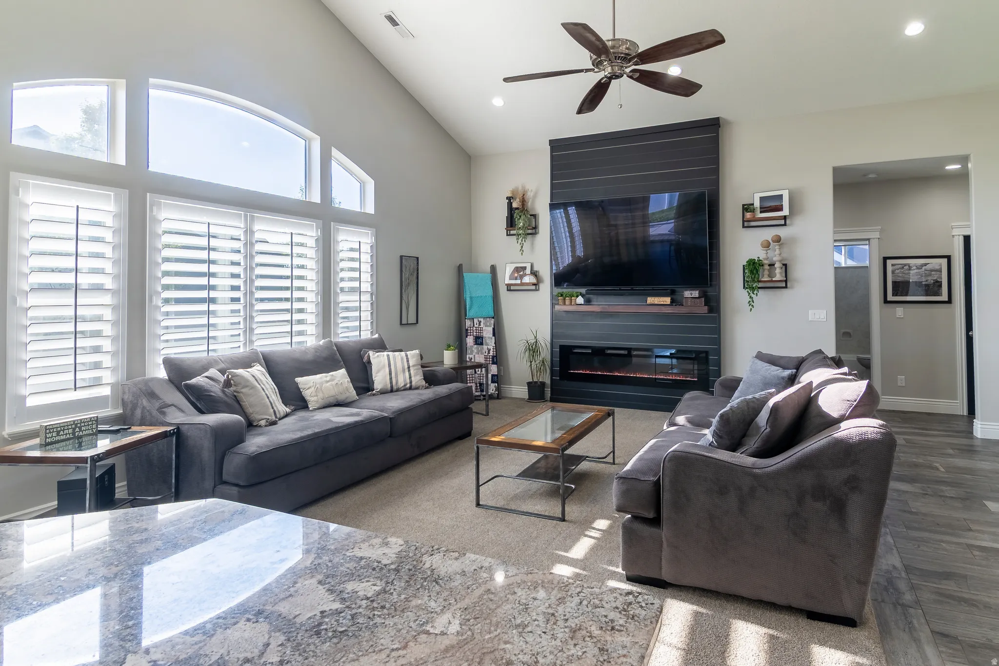 Vaulted ceiling living area featuring a prominent fireplace surround with built-in shelving, wood-finish flooring, a ceiling fan, and large windows with integrated plantation shutters