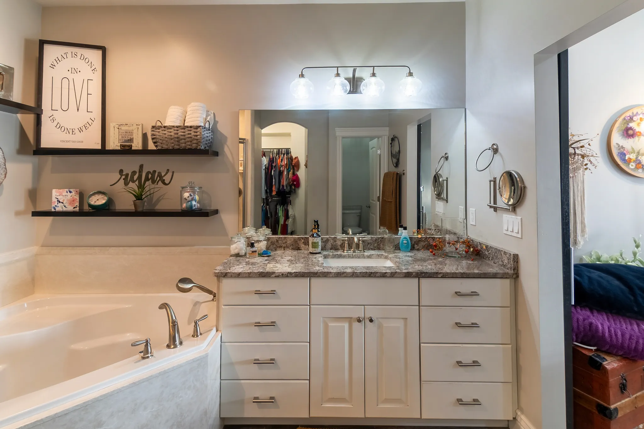 Bathroom featuring a built-in corner bathtub with deck-mounted faucet, a vanity with under-mount sink and brushed nickel faucet, a large wall-mounted mirror, a three-light vanity fixture, and open shelving