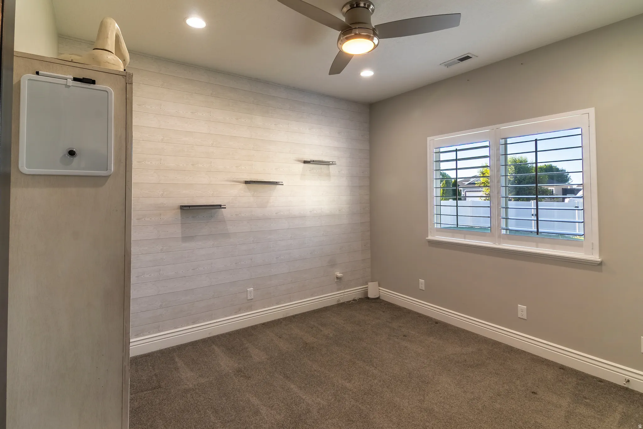 Carpeted room featuring a shiplap accent wall with integrated shelving, a ceiling fan with light fixture, recessed lighting, and a window with plantation shutters