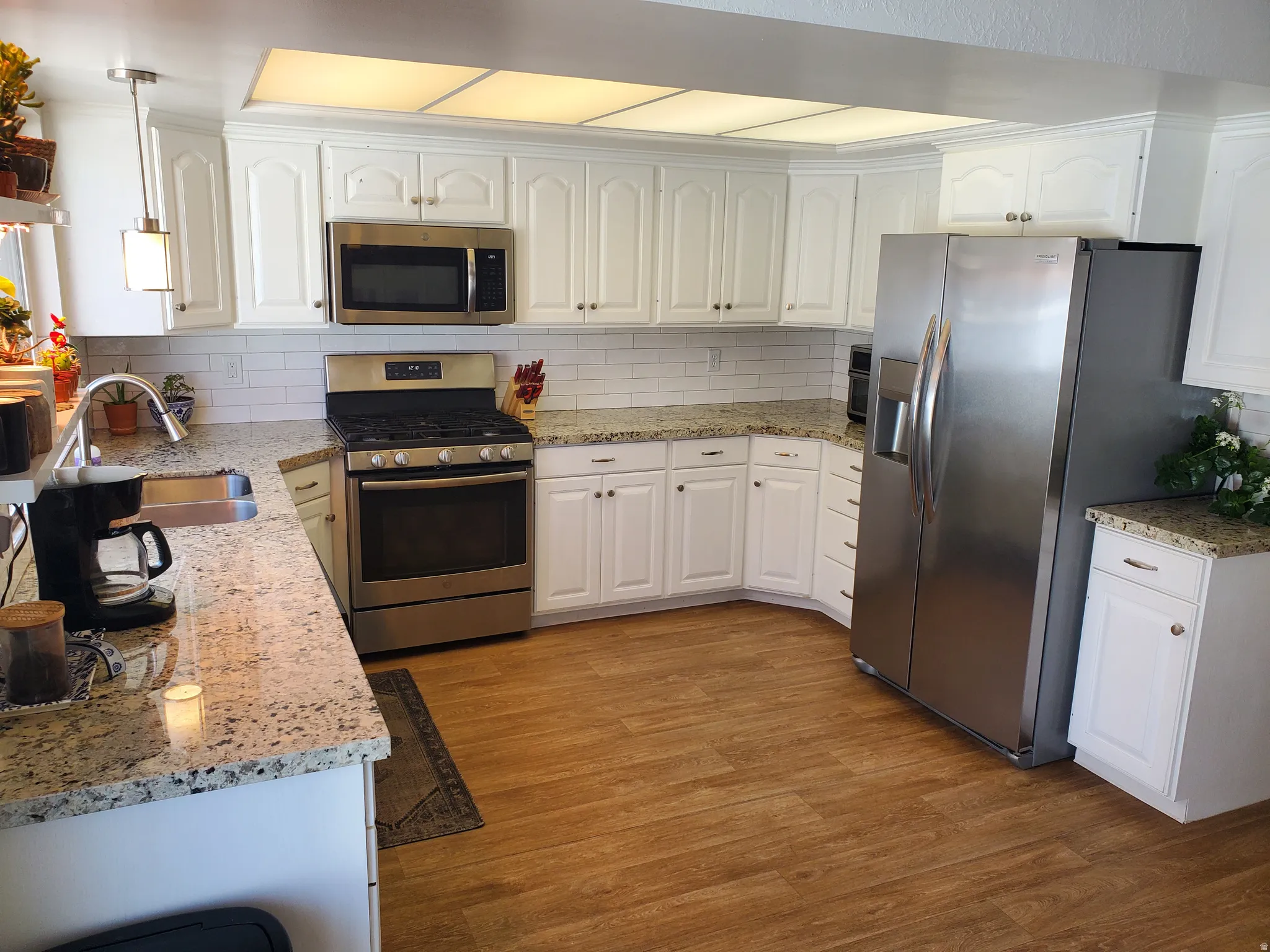 Contemporary kitchen featuring white cabinetry, light-toned granite countertops, a subway tile backsplash, stainless steel appliances, and wood-finish flooring