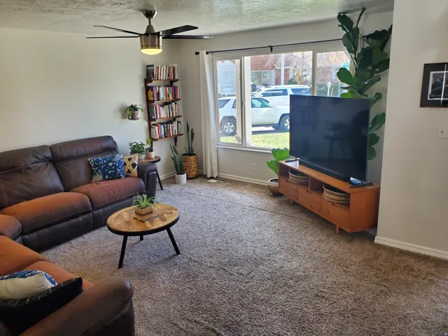 Living area featuring light beige walls, a ceiling fan with integrated lighting, and a large window with white blinds