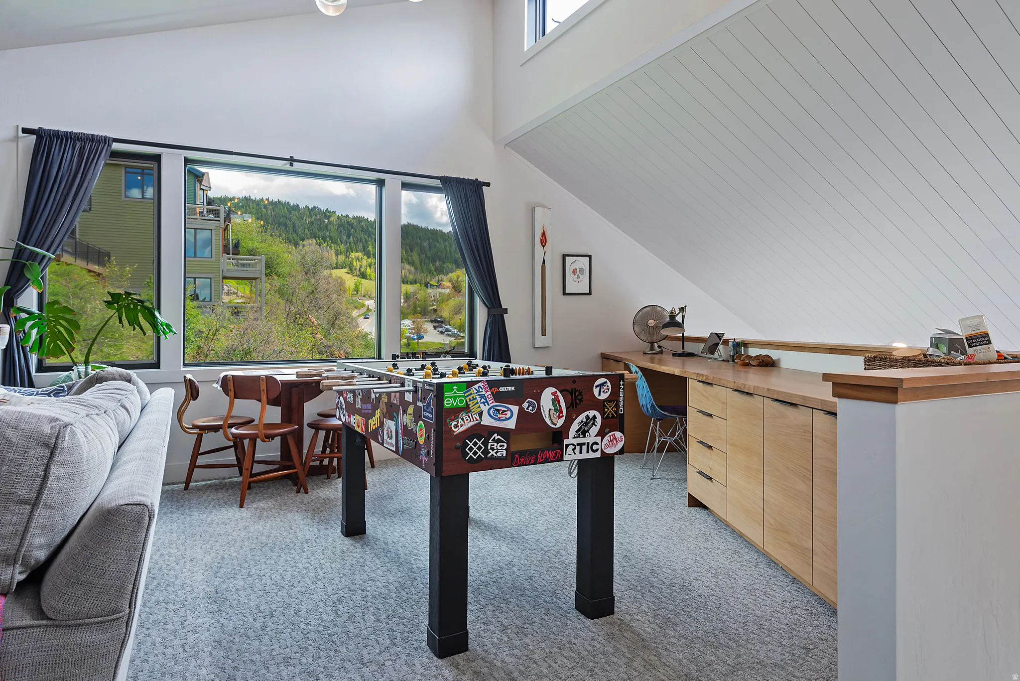 Expansive window wall with black trim, built-in desk with wood-finish cabinetry, vaulted ceiling with wood paneling, and light gray patterned carpeting
