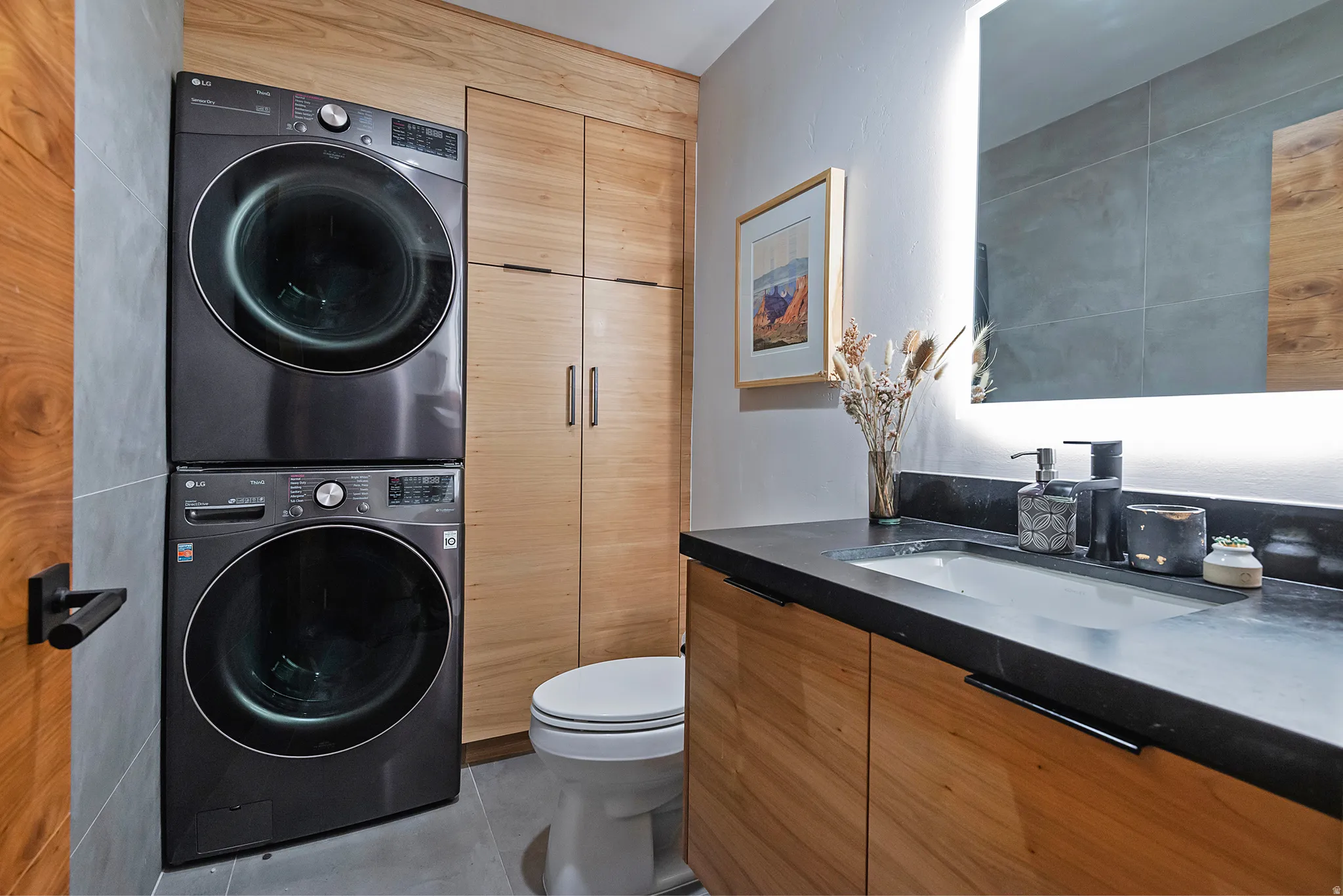 Compact laundry area with stacked front-load machines, a single-sink vanity with a dark countertop, wood-finish cabinetry, and an illuminated mirror
