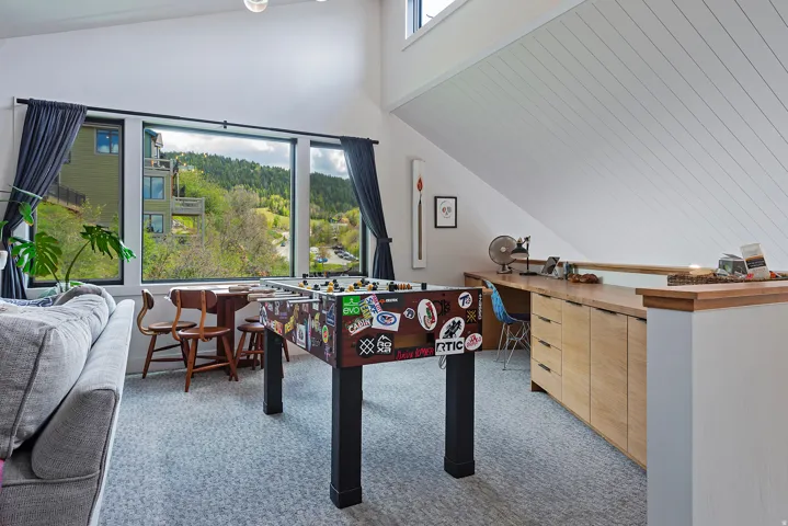 Expansive window wall with black trim, built-in desk with wood-finish cabinetry, vaulted ceiling with wood paneling, and light gray patterned carpeting