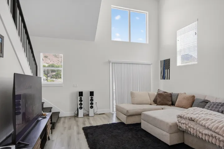 Spacious living area featuring high ceilings, white walls, wood-finish flooring, and a sliding glass door with vertical blinds