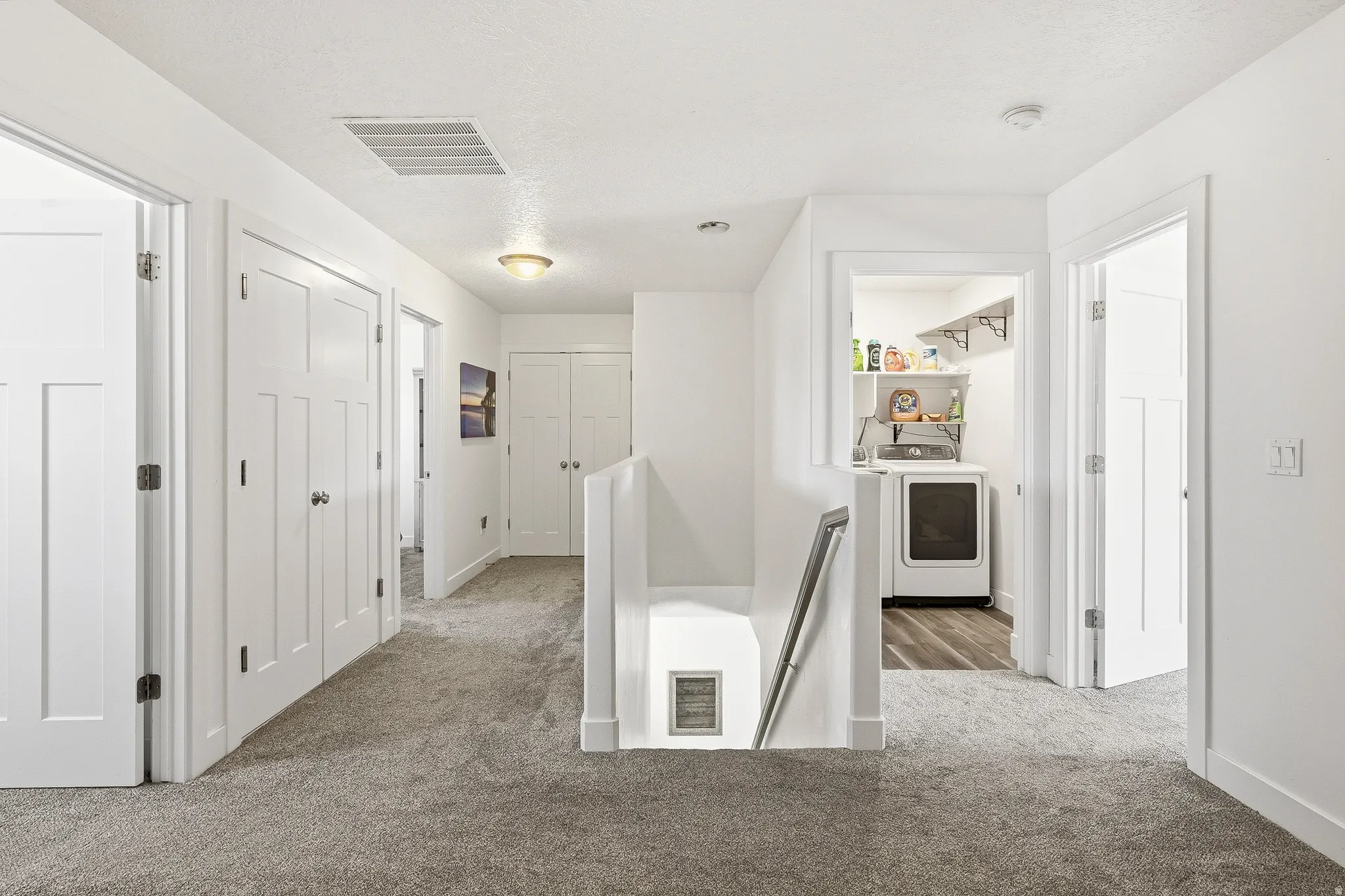 Hallway featuring light gray carpeting, white panel doors with black hardware, a laundry area with wood-finish flooring, and a staircase with a metal handrail