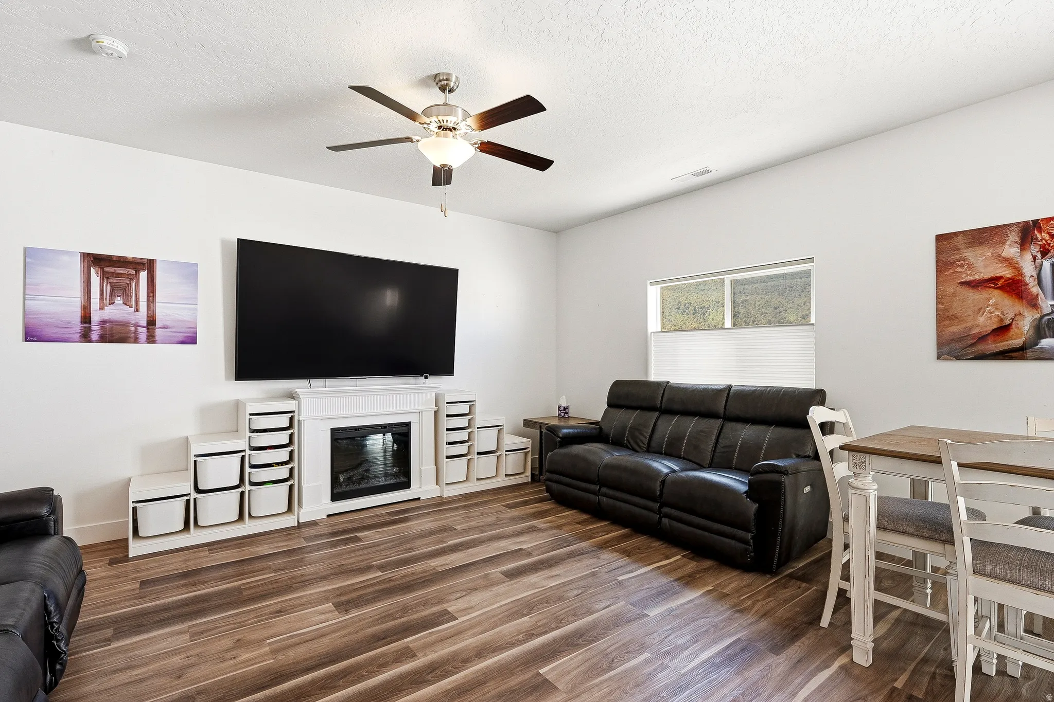 Living area featuring wood-finish flooring, a ceiling fan with integrated lighting, and a window with blinds