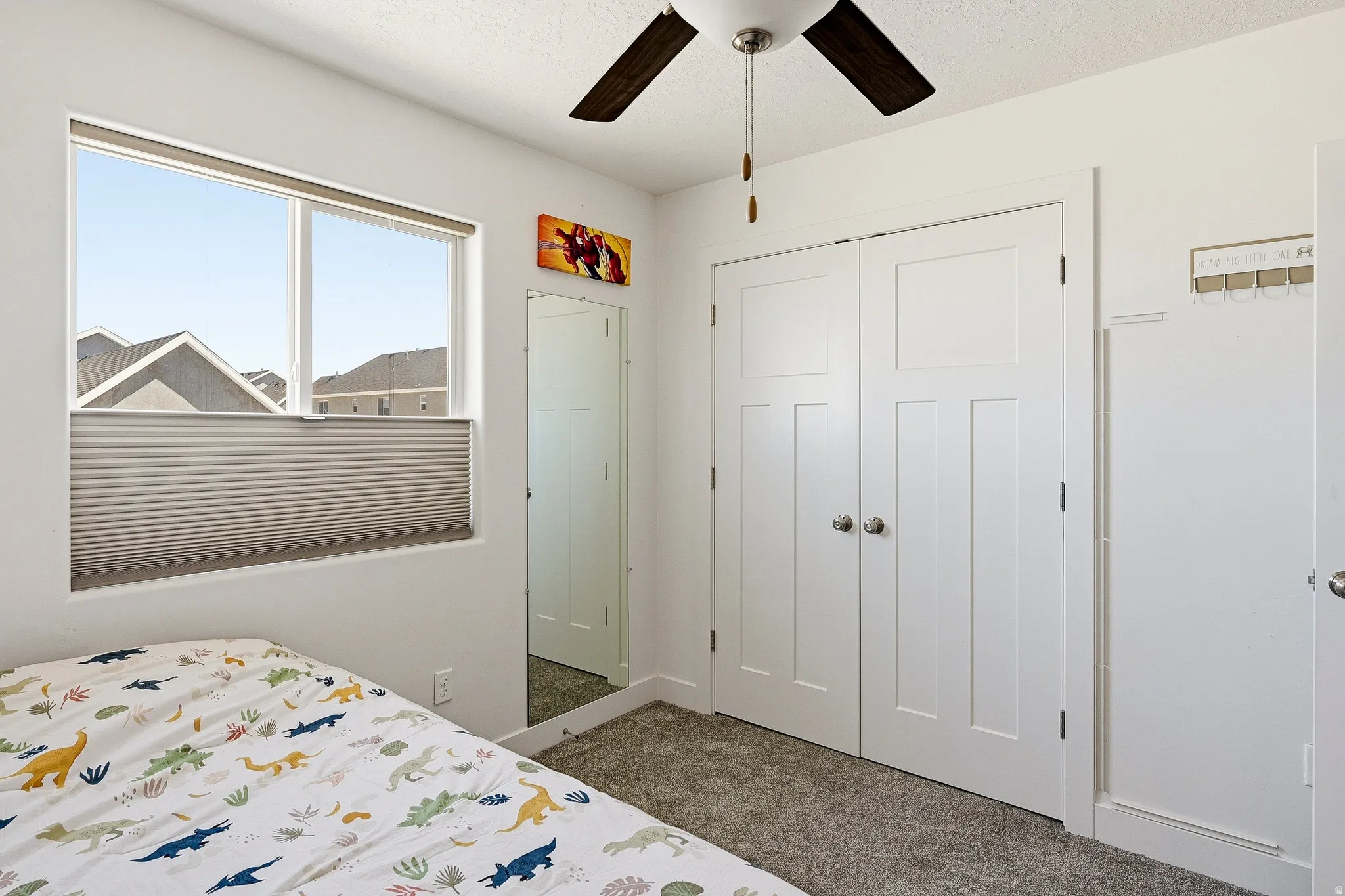 Bright room featuring a large window with privacy blinds, plush grey carpeting, a ceiling fan with wood-finish blades, and white bi-fold closet doors