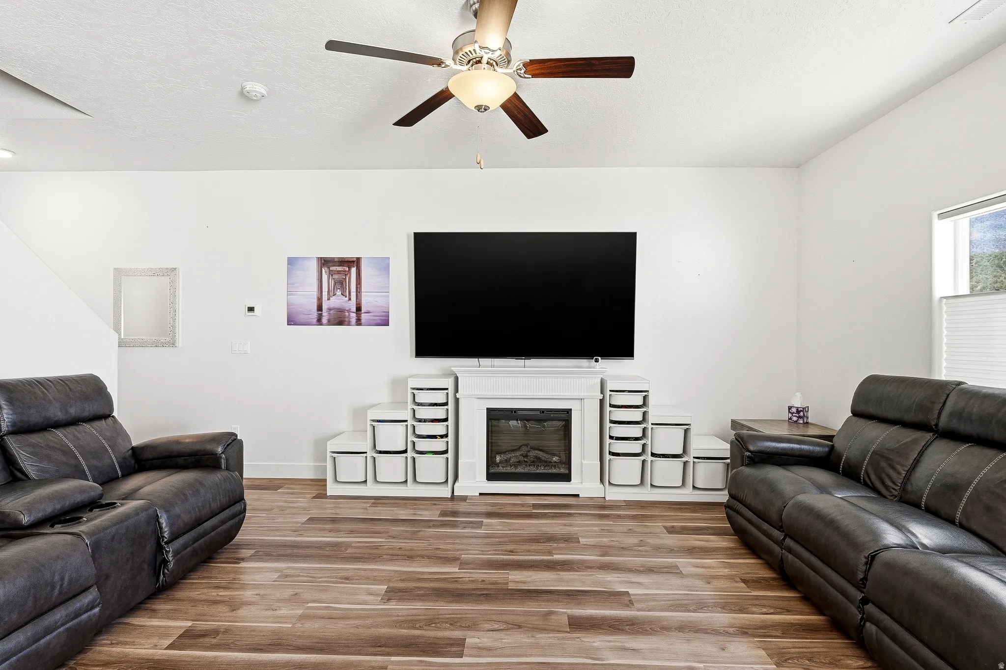 Spacious living area featuring wood-finish flooring, a ceiling fan with integrated lighting, and a white fireplace surround