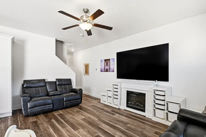 Living space featuring wood-finish flooring, a contemporary electric fireplace with white surround, built-in storage shelving, a ceiling fan with integrated lighting, and a visible staircase