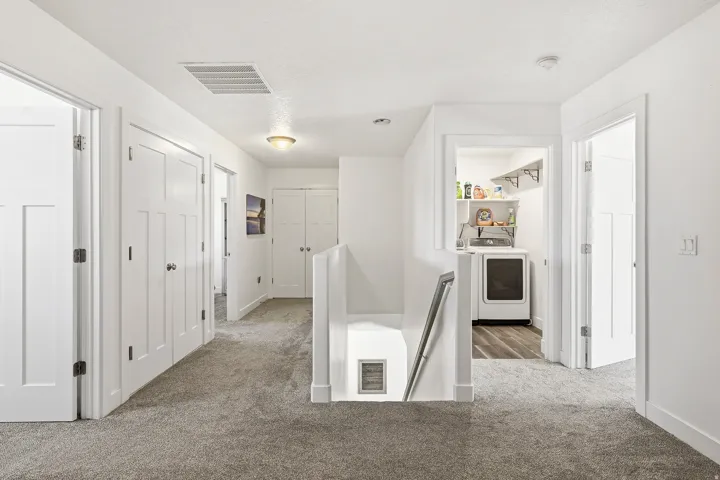 Hallway featuring light gray carpeting, white panel doors with black hardware, a laundry area with wood-finish flooring, and a staircase with a metal handrail