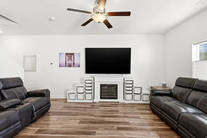 Spacious living area featuring wood-finish flooring, a ceiling fan with integrated lighting, and a white fireplace surround
