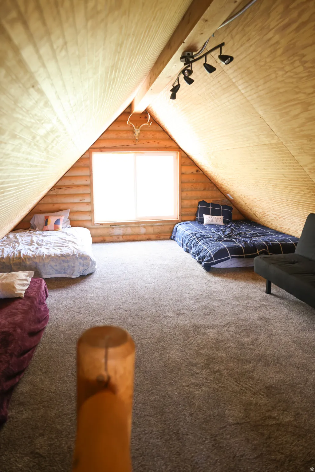 Spacious attic room featuring sloped wood-paneled ceilings, a bright central window, track lighting, and soft carpet flooring
