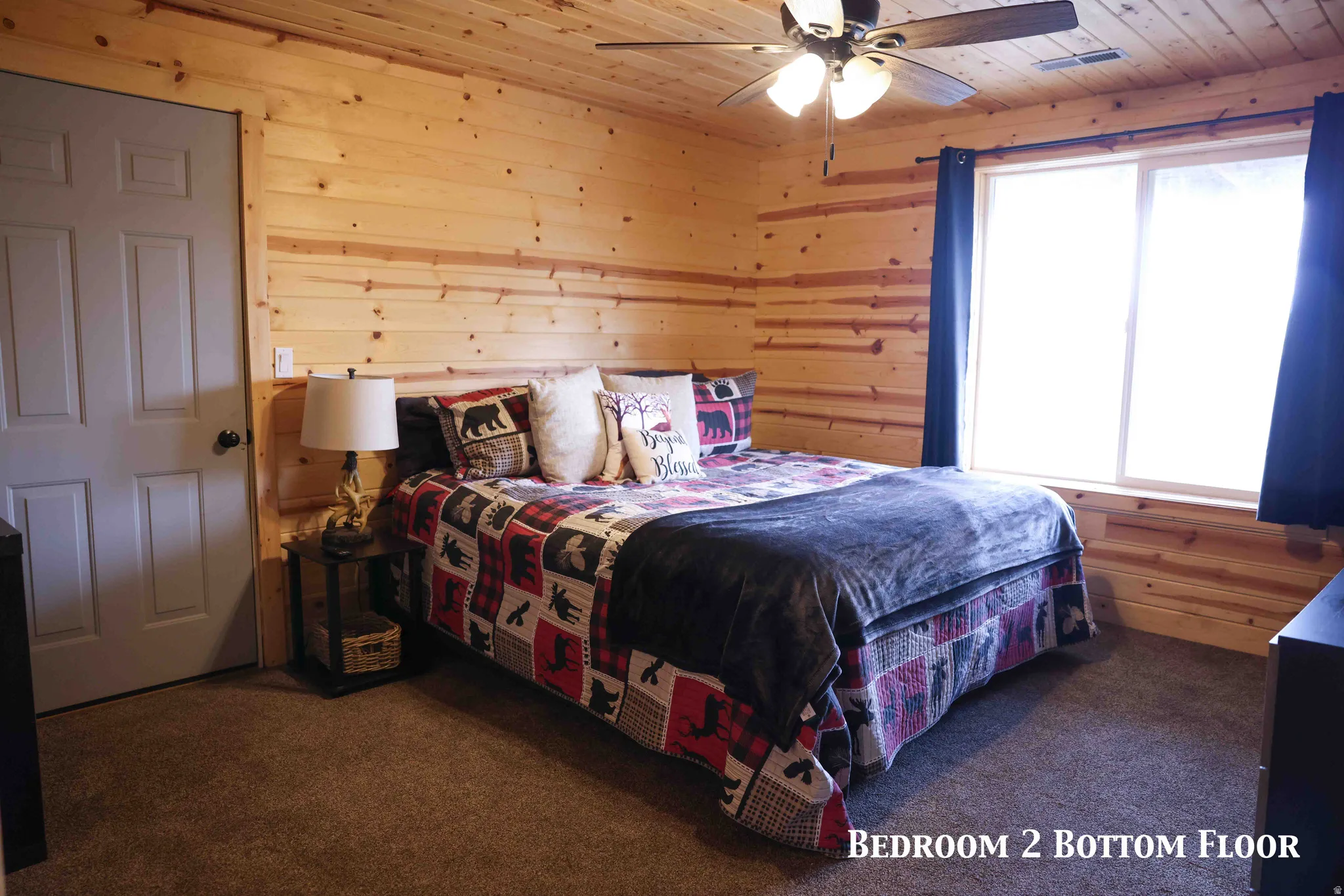 Pine wood paneling and ceiling throughout, featuring a ceiling fan with integrated lighting, a large window with privacy curtains, and soft carpet flooring