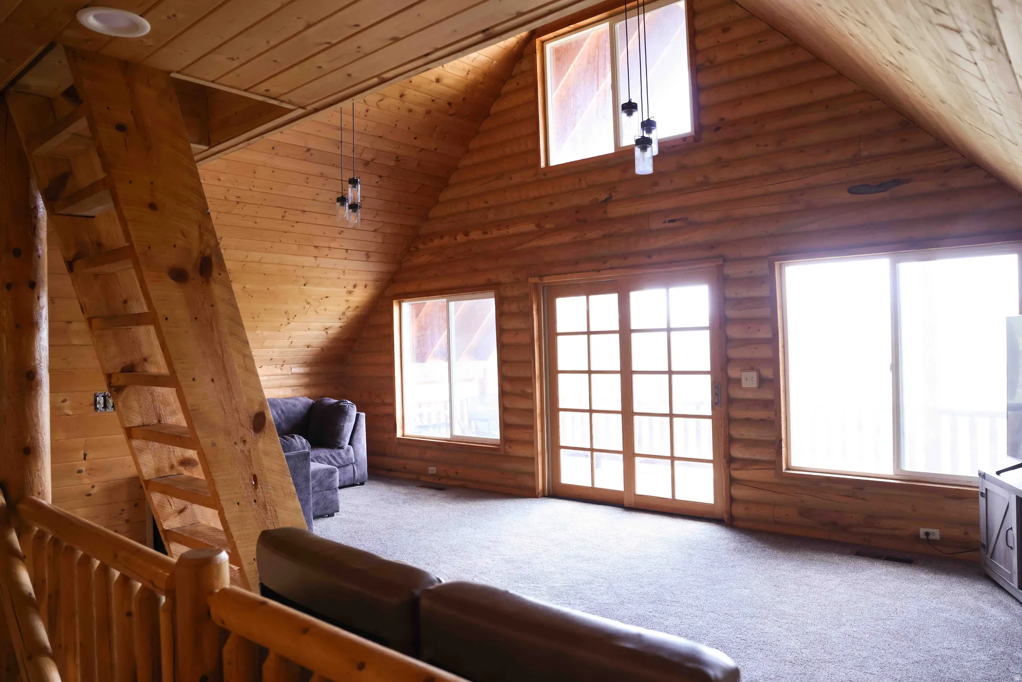 Vaulted ceiling great room featuring log wall construction, a wooden ladder-style staircase, two large windows, a glass-paneled double door, and a built-in recessed ceiling light