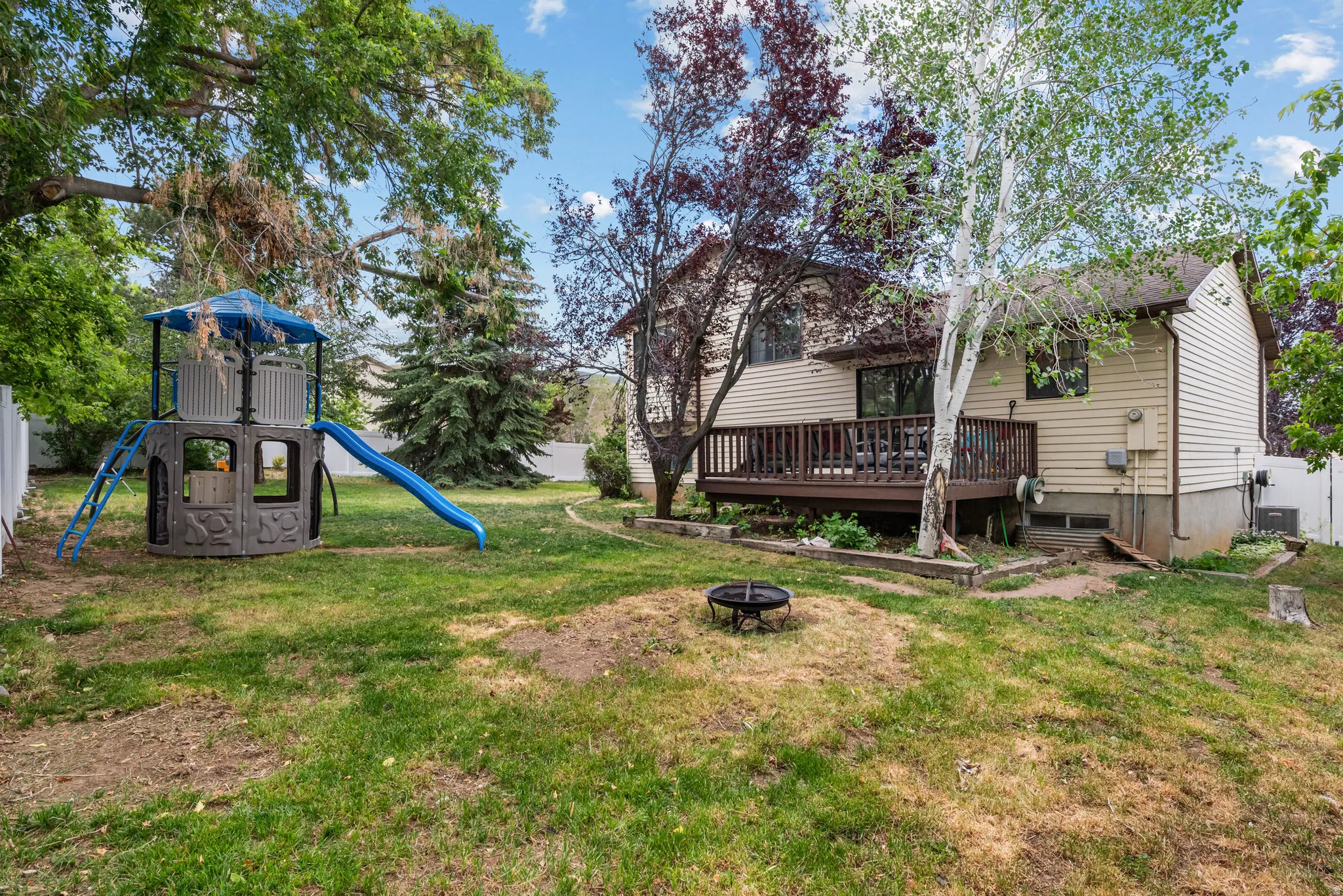 Expansive backyard featuring a multi-level deck, mature deciduous and evergreen trees, a fire pit area, and partial white fencing